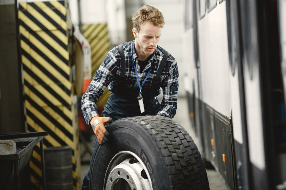 Mechanic lifts tire beside bus in garage.